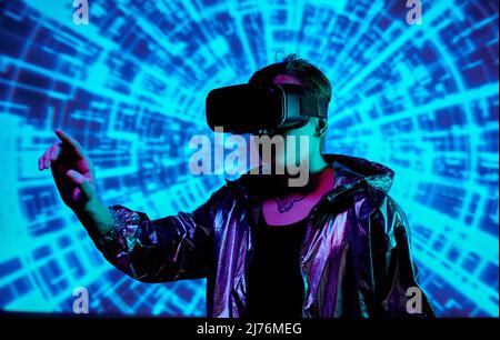 Man in vinyl hoodie jacket wearing virtual reality goggles pointing aside while traveling in virtual world Stock Photo
