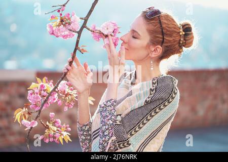 Young redhead woman in poncho smelling cherry blossom in park Stock Photo