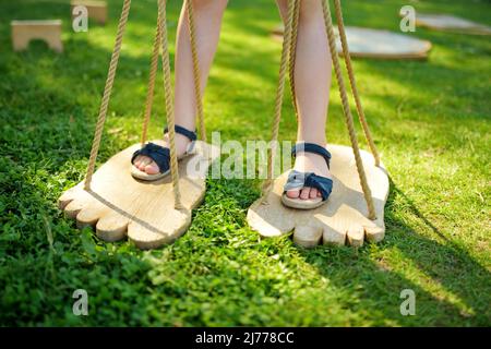 Little girl trying to balance walking on giant wooden feet in the park. Child playing funny outdoor game on sunny summer day. Family leisure for kids Stock Photo