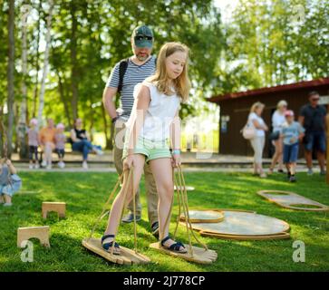 Little girl trying to balance walking on giant wooden feet in the park. Child playing funny outdoor game on sunny summer day. Family leisure for kids Stock Photo