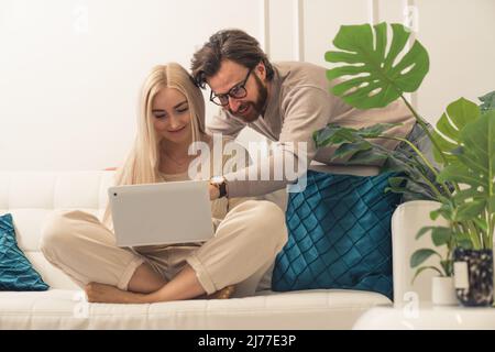 Blonde caucasian attractive woman sitting cross legged on a sofa with a laptop browsing internet, bearded man leaning towards her pointing at a screen. High quality photo Stock Photo