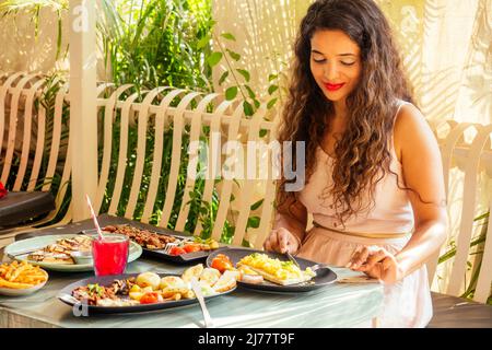 indian pretty woman in pink dress and curly hairstyle sitting in summer tropical cafe Stock Photo