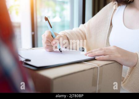 Close up Delivery man in red uniform holding package while client woman signing signature on paper receiving documents. Stock Photo