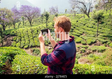 bloger redhaired ginger male enjoying morning taking selfie pictures on camera of smartphone in India chai plantations Munnar Stock Photo