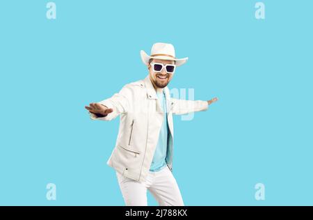 Happy young man in white leather jacket, cowboy hat and sunglasses dancing in studio Stock Photo