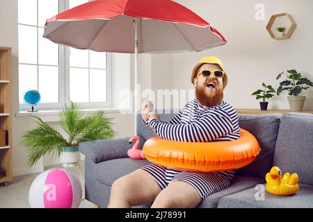 Funny chubby man having fun sitting on sofa in living room with inflatable swimming circle. Stock Photo