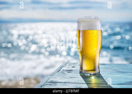 Cooled glass of beer with condensation drops on the blue wooden table. Blurred sea is at the background. Stock Photo