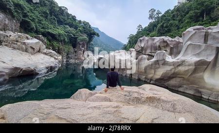 isolated young man sitting on naturally formed white shiny stone in unique shape at dry river bed image is taken at Sliang wah Umngot amkoi jaintia hi Stock Photo