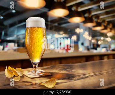 Cooled glass of beer with condensate on the wooden table. Nearby are potato chips. Blurred bar at the background. Stock Photo