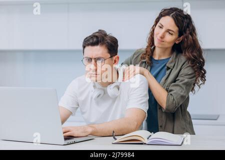 Wife is massaging the shoulders of her tired husband, a freelancer programmer in glasses and headphones, working on a laptop at home in the kitchen. Husband took a break from work. Stock Photo