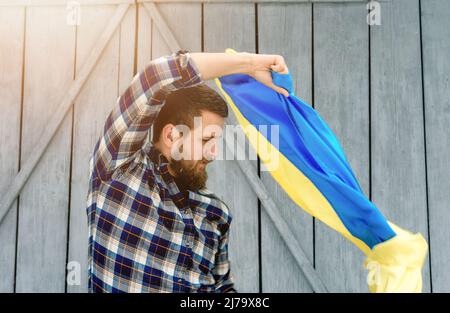 The man in a checkered shirt holds the state flag of Ukraine outdoors in snowy and sunny weather. Ukrainian flag, patriotism. Stock Photo