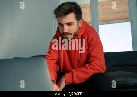 Young man wearing red sweatshirt watching movie on laptop in his room, hang out alone Stock Photo
