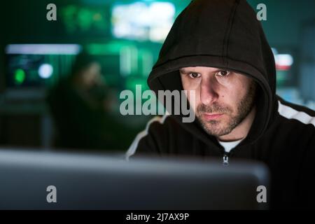 Portrait of mysterious hooded hacker sitting at desk and breaking into government data servers in dark atmosphere with system codes animation in Stock Photo