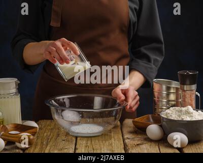 Cooking dough in a large glass bowl by the hands of a professional chef on a wooden kitchen table. Ingredients, many objects. Preparation of dough pro Stock Photo