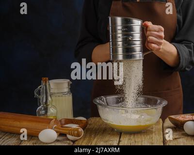 The chef prepares the dough in a glass bowl on a wooden kitchen table. Sifts flour through a sieve. Flour in frozen flight. Recipes made from dough. R Stock Photo
