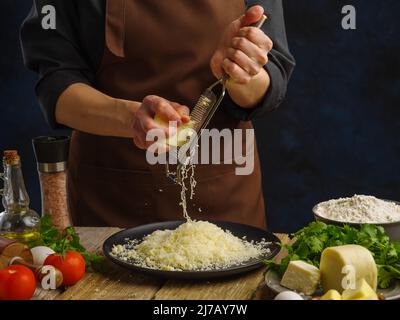 Cooking dishes from cheese, vegetables, herbs. Professional chef grates cheese on a wooden kitchen table, dark blue background. Cheese in a frozen fli Stock Photo