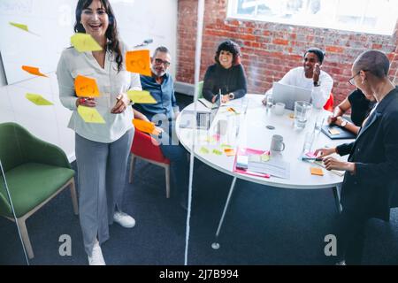 Smiling businesswoman making notes on a glass wall with her colleagues in the background. Creative young businesswoman sharing her ideas with her team Stock Photo