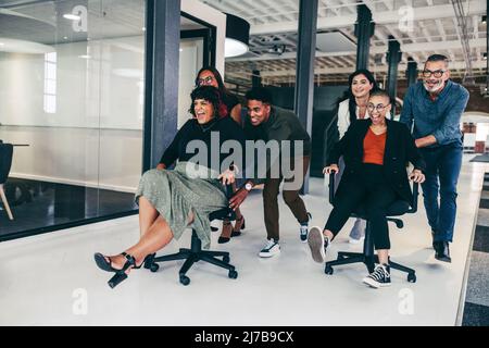 Fun during business hours. Cheerful team of businesspeople pushing each other on chairs in a modern office. Happy business colleagues having fun and c Stock Photo