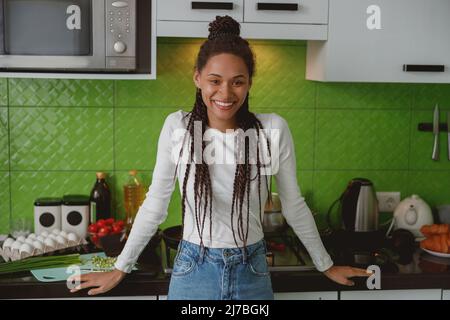 Portrait of cheerful young mixed-race woman smiling to camera standing in the kitchen Stock Photo