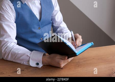 Caucasian businessman writing in notebook, male hands holding pen making notes planning new information appointments in personal paper planner on personal paper desk. A bearded guy in shirt and vest Stock Photo