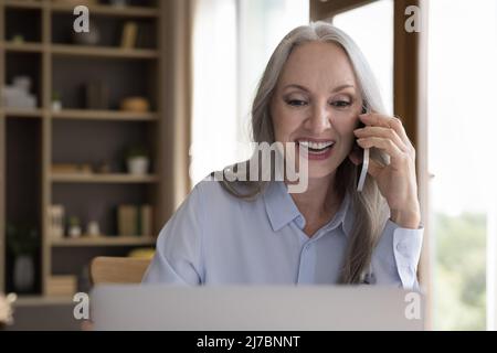 Happy 50s business lady talking on mobile phone at laptop Stock Photo