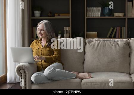 Happy senior laptop user woman sitting on couch at home Stock Photo