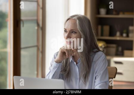 Pensive dreamy older 60s business lady sitting at laptop computer Stock Photo
