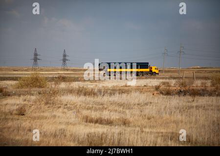 Transportation of large-diameter plastic pipes on yellow truck. Yellow dry grass in steppe. Grey sky. Stock Photo