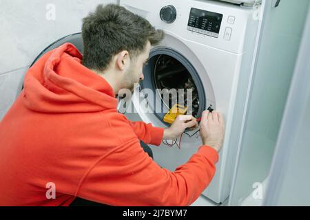 Repairman, young handsome man is repairing the broken washing machine in his house, he checks whether there is electricity with a voltmeter Stock Photo