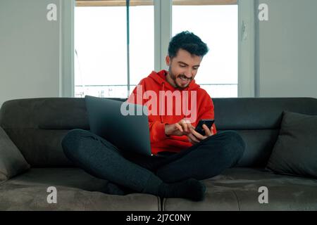 The young man sitting on the sofa has a laptop on his lap and is surfing the internet with his phone, young adult having a relaxed and enjoyable momen Stock Photo