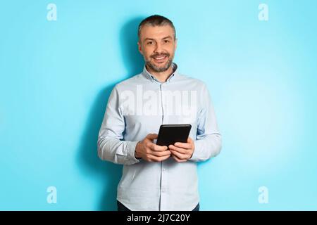 Middle Aged Businessman Holding Digital Tablet Standing Over Blue Background Stock Photo