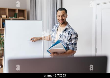 Cheerful arab male presenter having video conference via laptop and pointing at blank empty whiteboard, template mockup Stock Photo