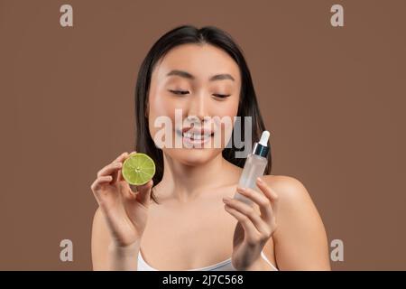 Fruit acids for skin care concept. Lovely asian lady holding lime and rejuvenating serum on brown studio background Stock Photo