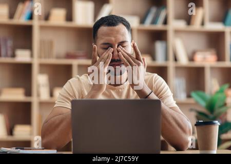 Tired arab freelancer guy sitting at workdesk in front of laptop at home and rubbing his eyes, suffering from burnout Stock Photo