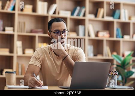 Focused arab man in glasses using laptop sitting at desk and writing in notebook, watching webinar studying online Stock Photo