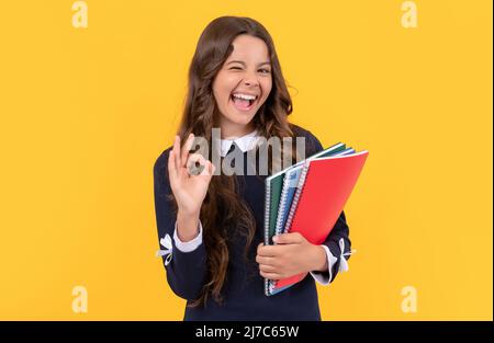 happy winking kid hold school copybooks studying on yellow background show ok gesture, school. Stock Photo