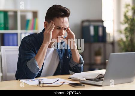 Stressed Young Male Entrepreneur Suffering Headache While Sitting At Desk In Office Stock Photo