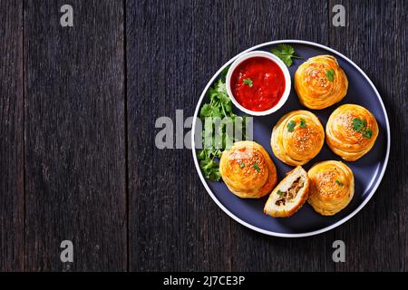 freshly baked snail shaped hand pies with ground mutton fillings with tomato sauce and fresh coriander on plate on dark wooden table, horizontal view Stock Photo