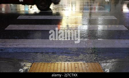 Rain drops on wet asphalt of city street in USA, raindrops falling on zebra crossroad. Cars lights reflection on road in rainy weather. Puddle of water by pavement. Night or twilight dusk atmosphere. Stock Photo