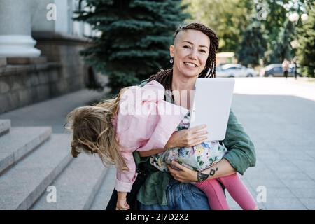 Freelance Parents. Busy working mom with baby toddler daughter on hands walk and looking on tablet in street. Multitasking Freelancer mom making notes Stock Photo