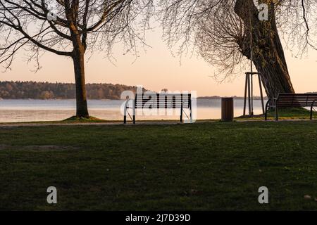 Empty park bench in front of an idyllic lake landscape. The sun is going down and the sky is glowing in orange color. Tranquil scenery without people. Stock Photo