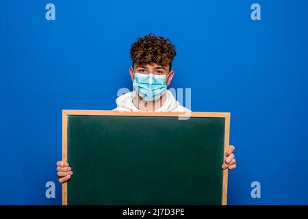 Young man with curly hair dressed in a white sweatshirt and surgical mask holding a blank white board, isolated on blue background Stock Photo