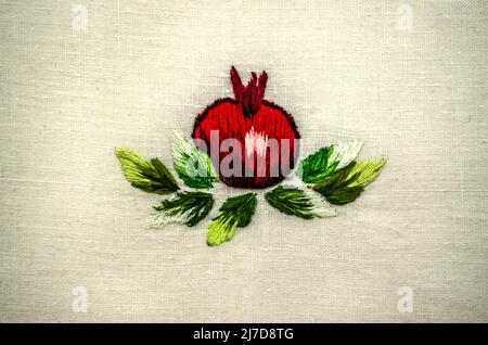 Yerevan, Armenia, April 04, 2022:Bright red round pomegranate embroidered with satin stitch among delicate curved autumn leaves with green tints on wh Stock Photo