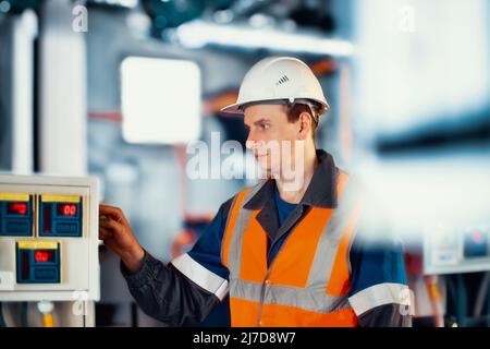 Operator or engineer in helmet and protective vest checks modern equipment in industrial plant. Portrait of Caucasian worker 30-40 years old working in production hall. Authentic scene workflow. Stock Photo