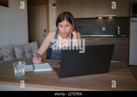 asian female freelancer working on laptop in living room. Focused lady studying distantly and making notes in copybook, listening to lecture. technologies for online education and self classes Stock Photo