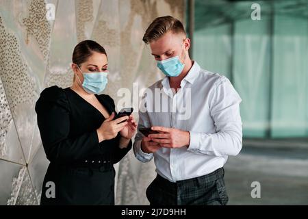 Coworkers with face masks using a mobile phone while talking about business outside the office building. Stock Photo