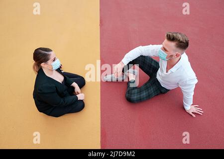 Business people wearing protective masks while sitting together on a different colored floor outdoors. Stock Photo