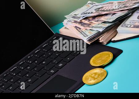 Black laptop with some bitcoins on it, a notebook with a wad of dollars on a blue table with a green background and copy space. Horizontal format phot Stock Photo