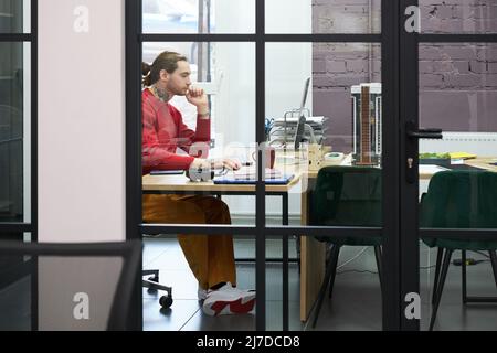 Young manager sitting at desk and working on computer behind the glass wall at modern office Stock Photo
