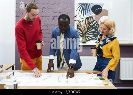 African man pointing at blueprint on table and discussing new project together with his colleagues Stock Photo
